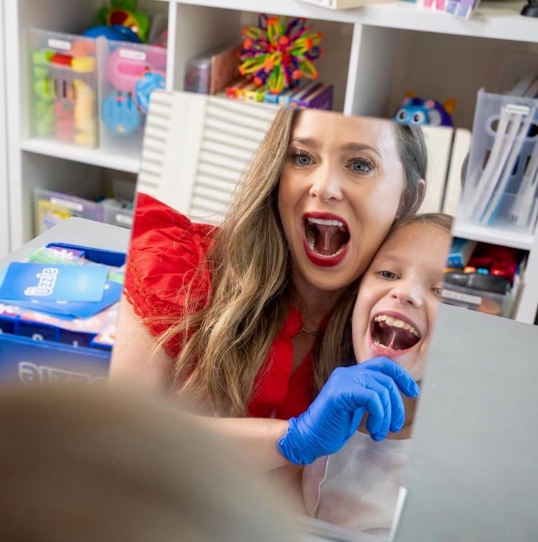 Laura and a young client practicing exaggerated mouth shapes in a mirror during myofunctional therapy